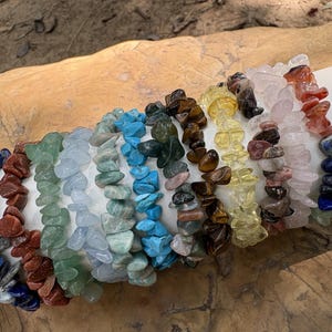 Puede incluir: Una colección de pulseras de virutas de piedras preciosas en varios colores, incluyendo azul, rojo, verde y amarillo. Las pulseras están ensartadas y se muestran sobre una superficie cilíndrica blanca. Las piedras tienen una forma natural e irregular.