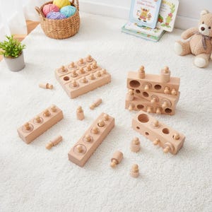 May include: A collection of wooden educational toys for children, including blocks with pegs and cylinders, arranged on a white rug. The toys are made of natural wood and are designed for learning and play.