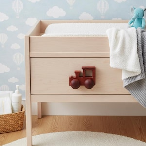May include: A light wood changing table with a red wooden train-shaped drawer pull. The table has a white changing pad, a white blanket, and a grey blanket. A blue stuffed elephant sits on top of the table.