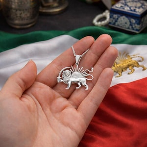 May include: A silver pendant necklace featuring a lion holding a sword, with a sunburst design. The pendant is displayed against a backdrop of a flag with red, white, and green stripes. The necklace is a piece of jewelry.