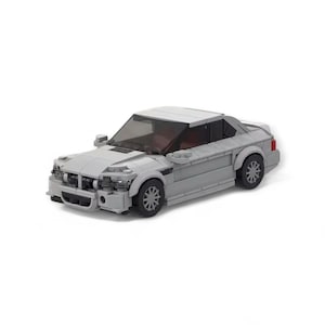 May include: A light grey toy car made from plastic bricks. The model has a detailed front grille, side mirrors, and black wheels. The car's windows are dark, and the interior appears to be red. The toy car is a coupe style.