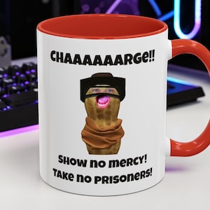 Puede incluir: Taza de cerámica blanca con interior y asa rojos. La taza presenta el texto "CHAAAAAARGE!!" sobre una imagen de un cacahuete con cara, con sombrero y bufanda. Debajo, las palabras "SHOW NO MERCY! TAKE NO PRISONERS!"