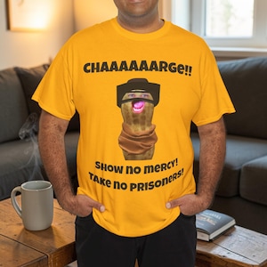 Puede incluir: Camiseta amarilla mostaza con un gráfico humorístico de una patata con sombrero de vaquero y bufanda. La camiseta tiene el texto "CHAAAAAARGE!!" encima de la patata y "Show no mercy! Take no prisoners!" debajo.