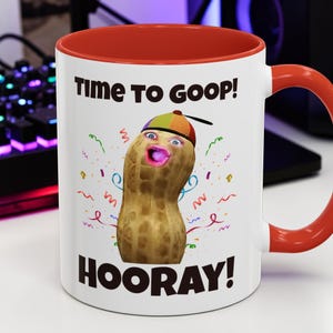 Puede incluir: Taza de cerámica blanca con interior y asa rojos. La taza presenta un personaje de cacahuete de dibujos animados con sombrero y las frases "TIME TO GOOP!" y "HOORAY!" en texto negro. Confeti colorido rodea el cacahuete.