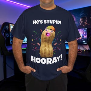 Puede incluir: Camiseta azul marino con un personaje de cacahuete de dibujos animados con un sombrero colorido y una boca rosa. El texto "He's STUPID! HOORAY!" está impreso encima y debajo de la ilustración del cacahuete.