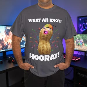Puede incluir: Una camiseta gris oscuro con un personaje de cacahuete de dibujos animados con un sombrero y una expresión de sorpresa. El texto "WHAT AN IDIOT!" y "HOORAY!" están impresos encima y debajo de la ilustración.