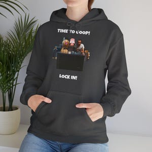 Puede incluir: Sudadera con capucha gris oscuro con bolsillo delantero y capucha con cordón. El gráfico en la parte delantera presenta un grupo de personas mirando una pantalla, con el texto "TIME TO LOOP!" arriba y "LOCK IN!" abajo.