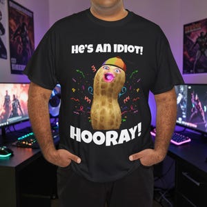 Puede incluir: Camiseta negra con el texto "He's an Idiot!" y "Hooray!". La camiseta presenta un personaje de cacahuete de dibujos animados con un sombrero arcoíris y una boca rosa. Confeti y serpentinas de colores rodean el cacahuete.