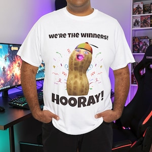 Puede incluir: Camiseta blanca con un cacahuete de dibujos animados con cara, gorro y boca rosa. La camiseta tiene el texto "WE'RE THE WINNERS!" y "HOORAY!" con confeti y líneas onduladas.