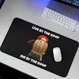 Puede incluir: Alfombrilla de ratón rectangular negra con el texto "LIVE BY THE GOOP DIE BY THE GOOP". La alfombrilla presenta un cacahuete de dibujos animados con una gorra roja y un martillo. Un ratón de ordenador blanco está a la derecha.