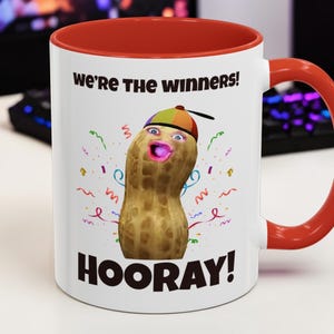 Puede incluir: Taza de cerámica blanca con interior y asa rojos. La taza presenta un personaje de cacahuete de dibujos animados con un sombrero, con el texto "We're the Winners!" y "HOORAY!" en negro. Confeti colorido rodea el cacahuete.