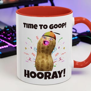 Puede incluir: Taza de cerámica blanca con interior y asa rojos. La taza presenta el texto "Time to Goop!" y "Hooray!" junto con un personaje de cacahuete de dibujos animados con sombrero y confeti.