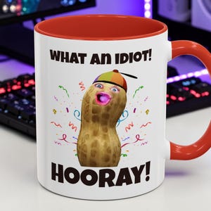 Puede incluir: Taza de cerámica blanca con interior y asa rojos. La taza presenta un personaje de cacahuete con cara, que lleva un sombrero, y las frases "WHAT AN IDIOT!" y "HOORAY!" en texto negro. Confeti colorido rodea el cacahuete.
