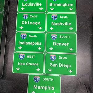 May include: A collection of green highway signs with white text and directional arrows. Signs include destinations like Louisville, Birmingham, Chicago, Nashville, Indianapolis, Denver, New Orleans, San Diego, and Memphis. Each sign features an interstate route number.