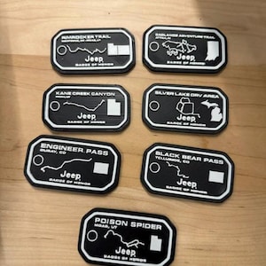 Peut inclure: Emblèmes Jeep Badge of Honor noirs et blancs. Chaque emblème présente le nom d'un sentier différent et un plan. Le texte "Jeep Badge of Honor" est inclus sur chaque emblème.