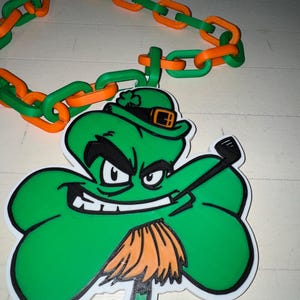 May include: A green shamrock pendant with a cartoon leprechaun design, featuring a black outline and a white border. The pendant is attached to a chain of alternating orange and green links, perfect for St. Patrick's Day.