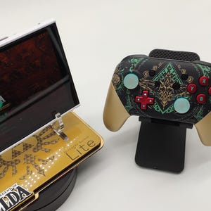 Peut inclure: Une console Nintendo Switch Lite et une manette, toutes deux sur le thème de The Legend of Zelda. La console est dorée avec des graphismes Zelda, et la manette est noire, dorée et sarcelle. L'écran de la console affiche un jeu Zelda.