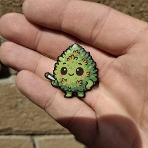 Puede incluir: Un pin de cogollo de cannabis de dibujos animados verde y amarillo con una cara sonriente y un pequeño porro. El pin tiene un contorno negro y se sostiene en una mano.