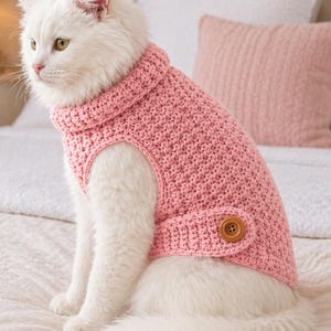 Könnte beinhalten: Eine weiße Katze trägt einen rosa gehäkelten Pullover mit hohem Kragen und einem einzelnen braunen Knopf an der Seite. Der Pullover hat ein strukturiertes Strickmuster und bedeckt den Oberkörper der Katze, während Beine und Schwanz frei bleiben.
