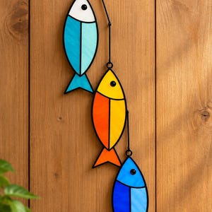 Puede incluir: Una decoración colgante vertical con tres peces estilizados hechos de vidrio tintado con marco de metal negro. El pez superior es blanco y azul claro, el del medio es amarillo y naranja, y el inferior es de dos tonos de azul.