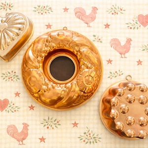 May include: Three antique copper molds in various shapes: a heart, a ring, and a round mold with a decorative pattern. The molds are displayed on a patterned background with rooster and heart motifs.