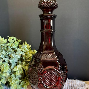 May include: A ruby red glass decanter with a textured stopper. The decanter has a geometric design with circular and diamond patterns. The bottle is approximately 10 inches tall.