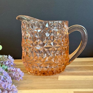 May include: A vintage peach-colored glass pitcher with a diamond pattern. The pitcher has a handle and a spout for pouring. The pitcher is sitting on a wooden surface with purple flowers in the background.