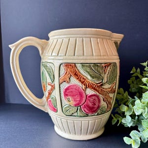 May include: A cream-colored ceramic pitcher featuring a raised, embossed design of red apples on brown branches with green leaves. The pitcher has a vertical ribbed pattern around the top and bottom sections and a curved handle on the left side.