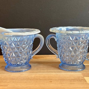 May include: Two light blue glass creamers with a diamond pattern and handles. One creamer has a spout. The rims are scalloped. The creamers are on a wooden surface.