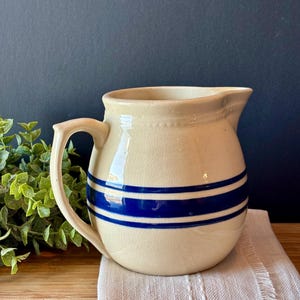 May include: A cream-colored ceramic pitcher featuring a glossy finish with a subtle crackle texture. The pitcher is decorated with two horizontal dark blue stripes encircling the body and includes a curved handle and a small pouring spout.