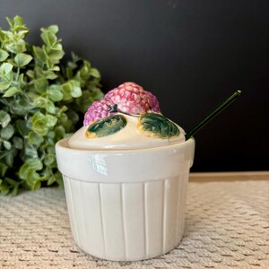 May include: A white ceramic lidded jar with vertical ridges on the base. The lid features a sculpted cluster of pink raspberries and green leaves. A thin, dark green spoon is inserted into the side of the jar, resting against the lid.