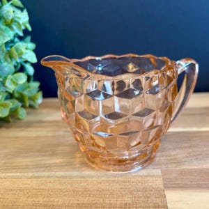 May include: A vintage peach-colored glass creamer with a geometric pattern. The creamer has a handle and a spout for easy pouring. The creamer is sitting on a wooden surface.