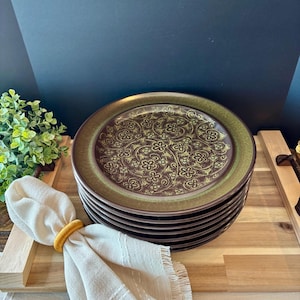 May include: A stack of brown and green ceramic plates with a floral design. The plates have a gold floral pattern in the center and a green border. A beige napkin with a yellow ring is in the foreground.