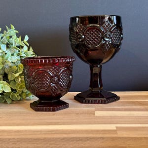May include: Two ruby red vintage goblets with ornate embossed patterns. One is a small tumbler, the other a stemmed wine glass. Both are made of glass and sit on a wooden surface. The background is dark.