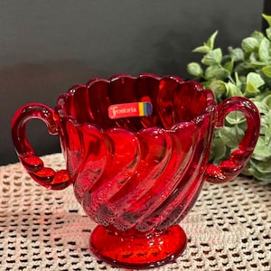May include: A vibrant red glass sugar bowl with two handles and a pedestal base. The bowl features a swirled design and a scalloped rim. The label "Fostoria" is visible on the side. This vintage piece is a decorative kitchen item.