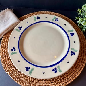 May include: A white ceramic plate with a blue border and berry design. The plate is decorated with blue berries and green leaves. The plate is placed on a woven placemat with a white napkin.