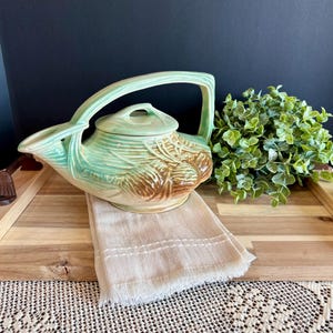 May include: A vintage ceramic teapot with a light green handle and lid, and a brown and green body. The teapot sits on a wooden tray with a beige linen towel. Decorative green foliage is also on the tray.