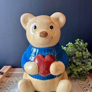 May include: A ceramic cookie jar shaped like a seated light tan bear. The bear wears a bright blue sweater with a ribbed collar and holds a large, glossy red heart against its chest. The bear has a smooth, glazed finish and a smiling face.