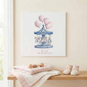 Op de afbeelding: Een canvas print met een aquarel illustratie van een carrousel met roze ballonnen. De carrousel is blauw en wit, met paarden. De print bevat de naam "ELLA" en de datum "15.8.2023". Een roze deken, rammelaar en babyslofjes liggen op een houten plank.