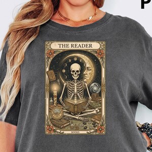 Op de afbeelding: Donkergrijs T-shirt met een tarotkaartontwerp met een skelet dat een boek leest, een maan en de tekst "THE READER". Het shirt heeft korte mouwen en een relaxte pasvorm.
