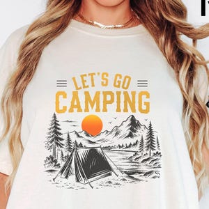 May include: An ivory short-sleeve t-shirt featuring a graphic print with the text "LET'S GO CAMPING" in yellow. The design includes a black and white illustration of a tent, pine trees, mountains, and a bright orange sun centered on the chest.