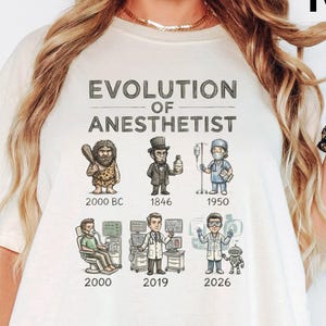 May include: An ivory short-sleeve t-shirt featuring a graphic titled Evolution of Anesthetist. The design displays six cartoon figures representing different eras from 2000 BC to 2026, including a caveperson, a historical figure, and medical professionals.