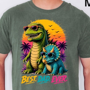 May include: A moss green t-shirt featuring a graphic of a large green dinosaur and a smaller blue triceratops, both wearing sunglasses. The background shows a yellow and orange sunset with palm trees. The text BEST. DAD. EVER. is printed below.