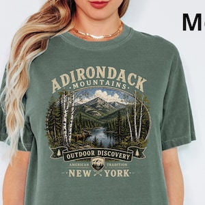 Puede incluir: Camiseta verde musgo con un gráfico de las montañas Adirondack y el texto "ADIRONDACK MOUNTAINS OUTDOOR DISCOVERY AMERICAN TRADITION NEW YORK". La camiseta tiene mangas cortas y cuello redondo.
