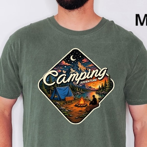 May include: A moss green short-sleeve t-shirt featuring a diamond-shaped graphic on the chest. The graphic depicts a camping scene with a blue tent, a campfire, mountains, and trees under a starry sky, with the text Camping Adventure.