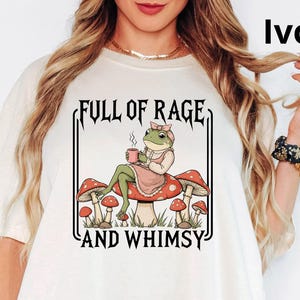 May include: Ivory-colored t-shirt with a whimsical graphic. The graphic features a frog in a dress and a bow, sitting on a mushroom and holding a mug. The text "FULL OF RAGE AND WHIMSY" is printed above and below the frog illustration.