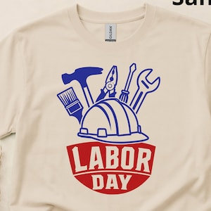 May include: A sand-coloured t-shirt featuring a graphic print of a blue hard hat surrounded by various tools, including a hammer, pliers, screwdriver, and wrench. Below the tools, the text "LABOR DAY" is printed in bold red capital letters.