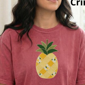 May include: Crimson-coloured t-shirt with a detailed pineapple design. The pineapple has a patchwork pattern in yellow and white, with green leaves. The shirt is a short-sleeved, casual style.