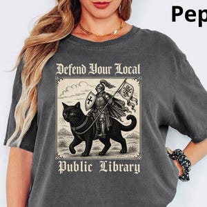 May include: Pepper-colored t-shirt featuring a graphic of a knight riding a black cat, with the text "Defend Your Local Public Library." The design is in a vintage style. The shirt has short sleeves and a crew neck.