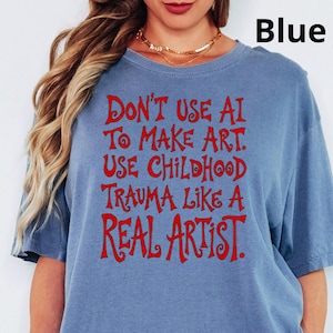 Może przedstawiać: Krótkorękawowy t-shirt w kolorze niebieskiego jeansu z czerwonym napisem: DON'T USE AI TO MAKE ART. USE CHILDHOOD TRAUMA LIKE A REAL ARTIST. Koszulka ma okrągły dekolt i luźny krój, a tekst jest wyśrodkowany na przedniej części klatki piersiowej.